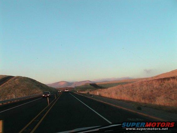 Highway scene with a dark sedan and a white car driving on a two-lane road with hills and a clear sky
