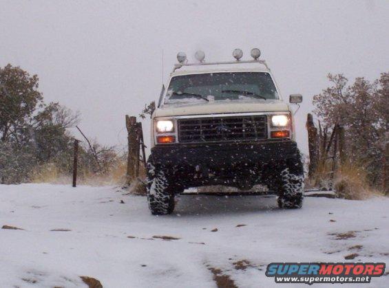 White SUV with off-road lights on roof driving in snow. Front grille visible