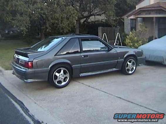 Gray 1987 Ford Mustang coupe with a rear spoiler and aftermarket wheels, parked on a driveway