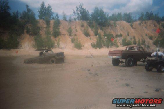 Two trucks stuck in mud, one partially submerged, with an ATV nearby on a sandy hill