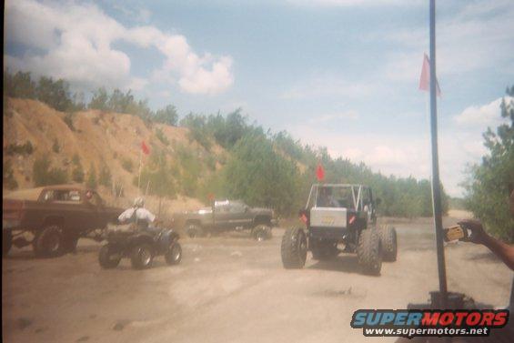 Several off-road vehicles and ATVs are parked on a dirt path with a sandy hill in the background