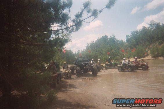 Group of ATVs and a Jeep on a muddy trail with trees and flags