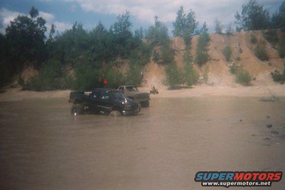 Two pickup trucks driving through shallow muddy water with trees and a sandy bank in the background