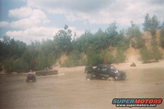 Black pickup truck and ATVs driving through muddy water near a sandy bank with trees