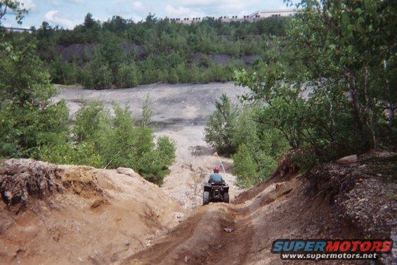 Person riding an ATV down a steep dirt path in a wooded, quarry-like area