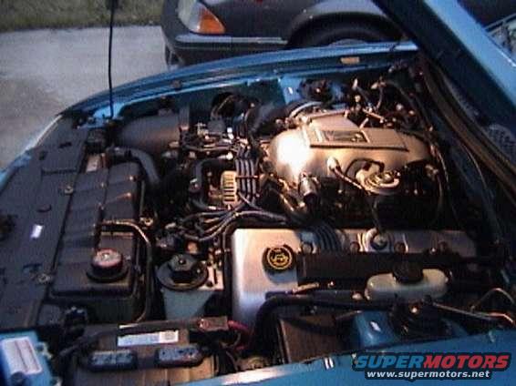 Engine bay of a blue car, showing the motor and various hoses and components. Another car is partially visible in the background