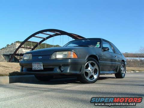 Dark gray coupe parked on asphalt, front 3/4 view, with a bridge and blue sky. License plate R11-DZP