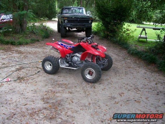 Red ATV with Confederate flag seat parked on dirt road, older truck behind it