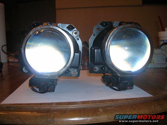 Pair of projector headlights with clear lenses and black housings, resting on a white surface