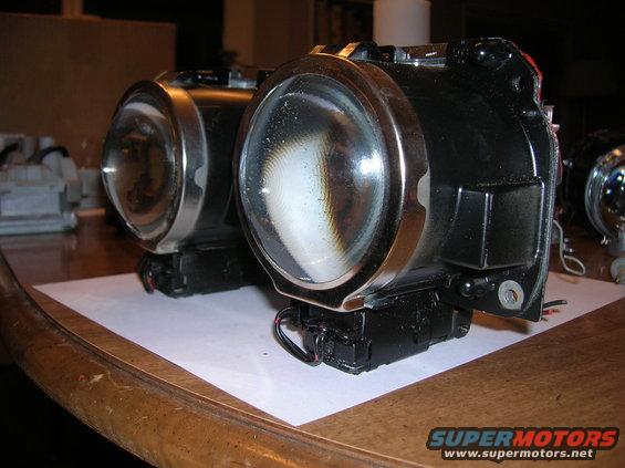 Two black headlight assemblies with chrome trim and clear lenses sit on a wooden table