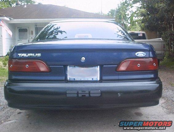 Rear view of a blue 1993 Ford Taurus parked in front of a house. A pickup truck is visible in the background