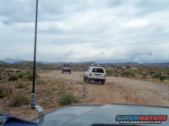 White 1991 Ford Bronco and red pickup truck drive on a dusty desert road under a cloudy sky