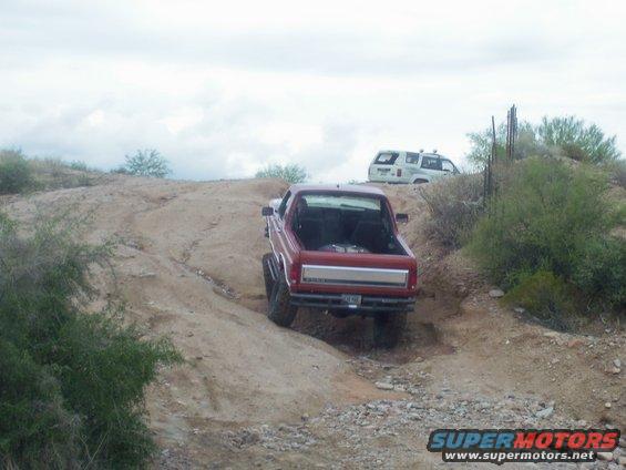 Red pickup truck with lifted suspension driving on a dirt trail, white SUV in background