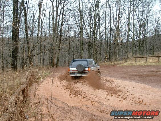 Ford Bronco driving through mud, rear view. Two-tone paint, spare tire on back