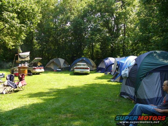 Camping scene with tents and two cars parked on grass, trees in background