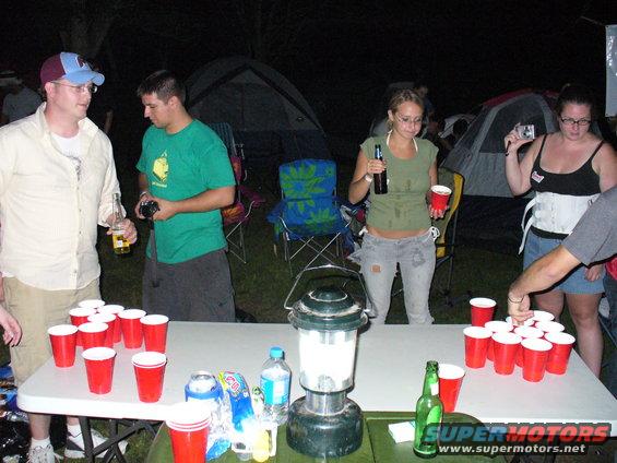 People gathered around a table with red cups set up for beer pong at a campsite at night