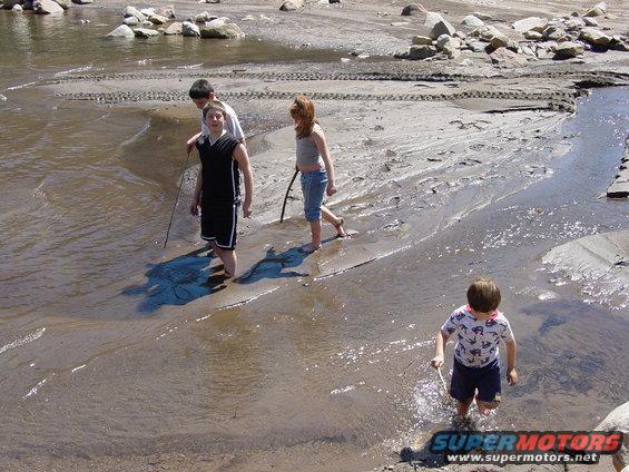 Four children wading in shallow water and mud near rocks and tire tracks