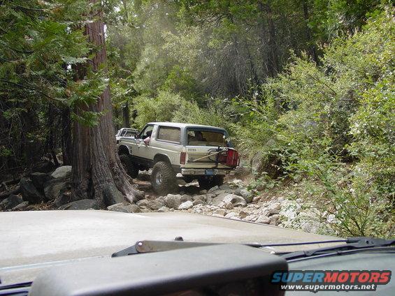 White Ford Bronco on a rocky trail, rear-mounted spare tire and fuel can visible