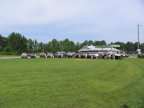 Several lifted trucks and SUVs parked in a grassy field near a gas station