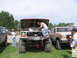 1995 Ford C-350 (Centurion Conversions) - A man works on the engine of a heavily modified red 1979 Ford F-Series truck, parked alongside other vehicles on a grassy field.