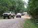 1995 Ford C-350 (Centurion Conversions) - A convoy of modified off-road vehicles travels down a wooded gravel road.