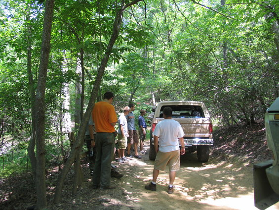 Group of people standing by a tan SUV on a dirt road in a wooded area. Another vehicle is partially visible on the right