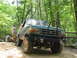 1995 Ford C-350 (Centurion Conversions) - A modified Ford Bronco sits on a rocky trail surrounded by lush green trees.