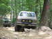 1995 Ford C-350 (Centurion Conversions) - A tan Ford Bronco drives over rocks on a wooded trail, with another vehicle visible behind it.