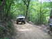 1995 Ford C-350 (Centurion Conversions) - A white pickup truck navigates a wooded trail, with another vehicle partially visible behind it.