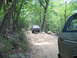 1995 Ford C-350 (Centurion Conversions) - A dark Ford F-Series truck navigates a rocky dirt path through a wooded area, with another vehicle visible in the foreground.