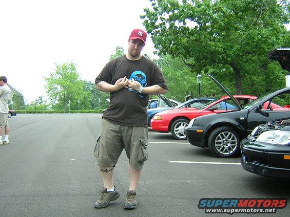 Guy in red hat and cargo shorts flexing in front of parked cars. One car is red, two are black