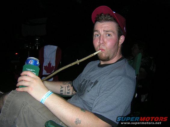 Man in a red cap and grey t-shirt holds a can and a stick in his mouth, with a Canadian flag chair behind him