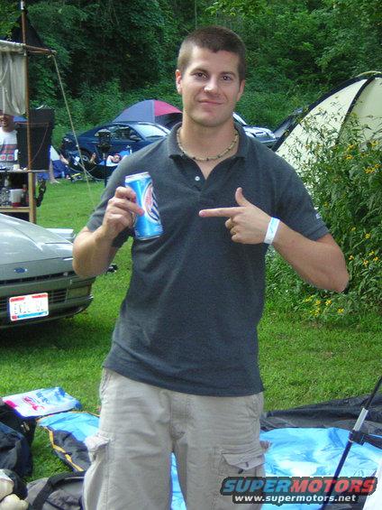 Guy with a can points at himself. A car is partially visible in front. Tents and trees in background