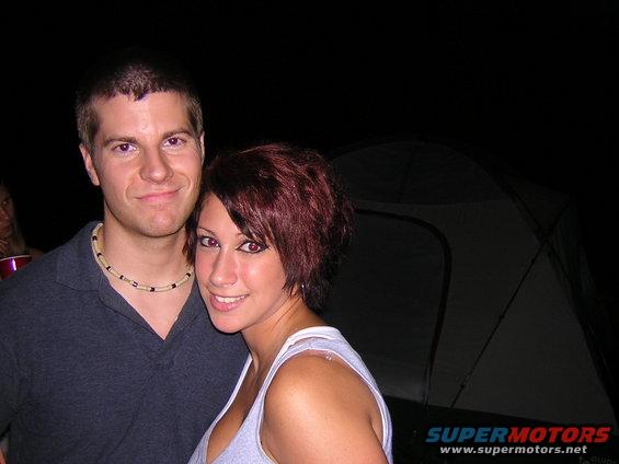 Guy and girl posing for a photo at night, with a tent behind them. Supermotors logo in the corner