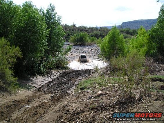 White SUV splashing through a muddy puddle in a dry riverbed with green trees and mountains in the background