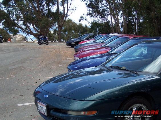 Several Ford Probes parked in a row, one in the foreground is dark green. A motorcycle is parked behind them