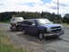 1955 Ford F-100 - A white Ford Bronco is transported on a trailer behind a blue pickup truck on a roadside.