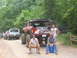 1995 Ford C-350 (Centurion Conversions) - A group of men and modified pickup trucks are gathered on a dirt road in a wooded area.