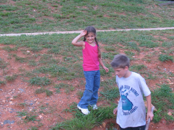 Girl in pink tank top and jeans waves, boy in t-shirt stands nearby in a dirt and grass area