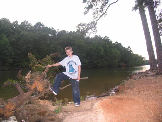 Boy in a t-shirt and jeans stands on a fallen tree by a lake with trees in the background