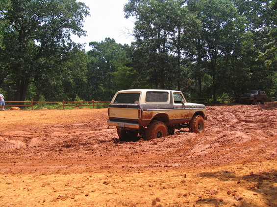 Tan and brown Ford Bronco stuck in mud, rear quarter view. Muddy tires and body. Another vehicle visible in background