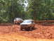 1995 Ford C-350 (Centurion Conversions) - Two SUVs navigate a muddy off-road course with trees and a wooden fence in the background.
