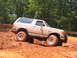 1995 Ford C-350 (Centurion Conversions) - A tan Ford Bronco is shown struggling through a muddy off-road trail, with trees and spectators in the background.