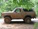 1995 Ford C-350 (Centurion Conversions) - A mud-covered tan Ford Bronco is seen from the side, parked on a dirt road under trees.