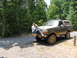 1995 Ford C-350 (Centurion Conversions) - A person sits on the custom bumper of a lifted Ford Bronco parked on a dirt road next to a wooded area.