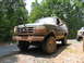1995 Ford C-350 (Centurion Conversions) - Mud-covered green Ford Bronco parked on a gravel shoulder next to a paved road, with a second truck in the background.