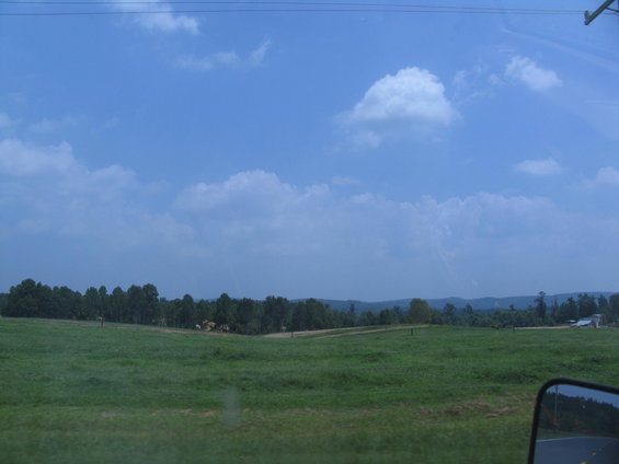 Side mirror view of a rural landscape with green fields and trees under a blue sky