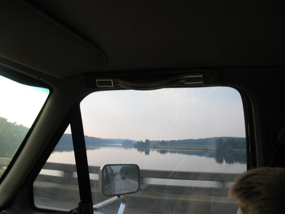 Interior view from a vehicle, looking out the passenger side window at a wide river and distant tree line. A side mirror reflects a road ahead