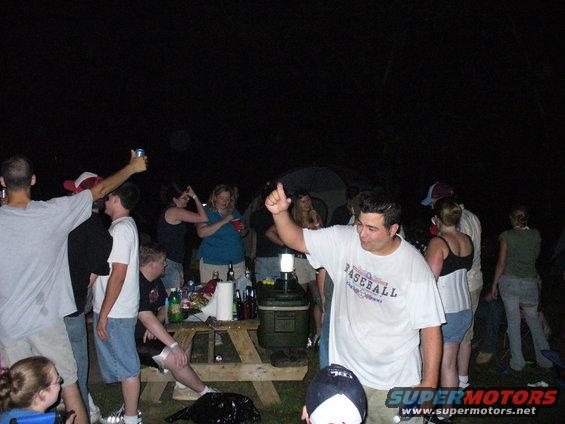 People gathered outdoors at night around a picnic table with drinks and a cooler