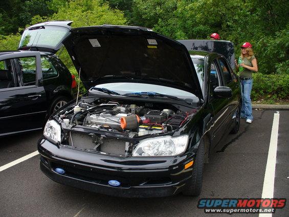 Black sedan with hood open, showing engine bay. Another black car is parked to the left. Two people in red hats are visible